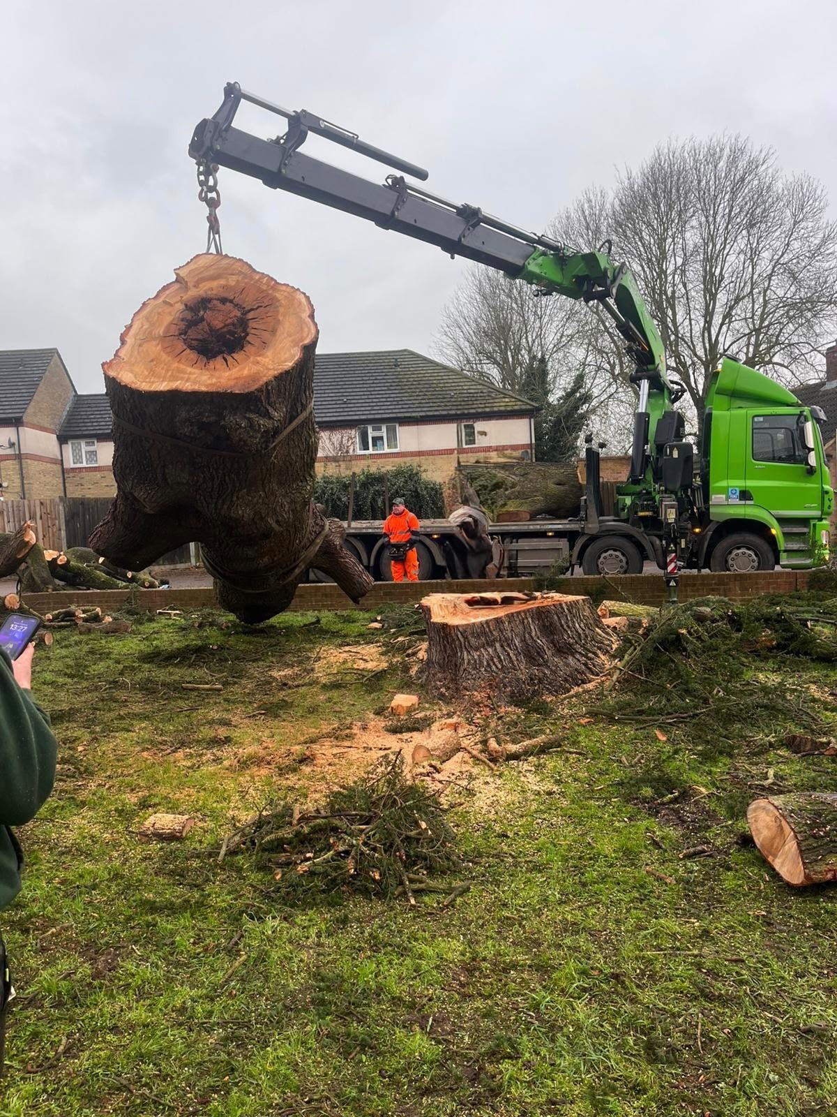 Vehicle-mounted crane lifting a large hollow trunk section clear of the ground-level stump during an urgent tree removal